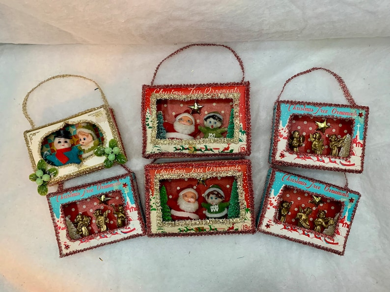 May include: Six vintage Christmas tree ornaments in the shape of small boxes with a window. Each box has a different scene inside, including Santa Claus, elves, and angels. The boxes are decorated with red, white, and blue paper and glitter.