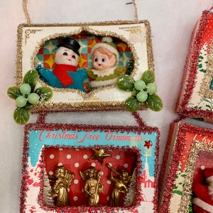 May include: Three vintage Christmas tree ornaments in boxes. The first ornament features a snowman and an elf in a winter scene. The second ornament features three gold angels in a winter scene with the text "Christmas Tree Ornaments". The third ornament features a Santa Claus in a red suit.