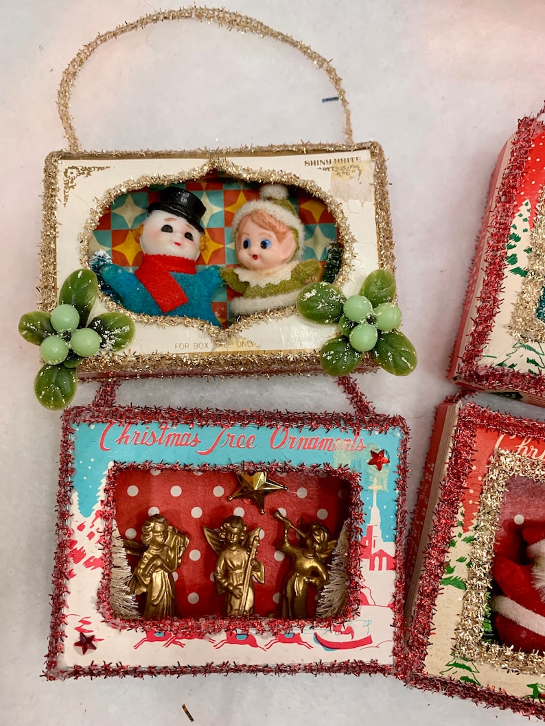 May include: Three vintage Christmas tree ornaments in boxes. The first ornament features a snowman and an elf in a winter scene. The second ornament features three gold angels in a winter scene with the text "Christmas Tree Ornaments". The third ornament features a Santa Claus in a red suit.
