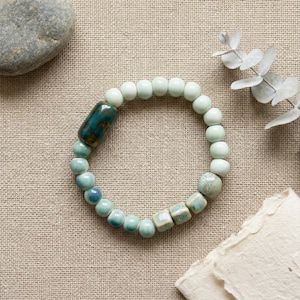 Könnte beinhalten: Ein Armband aus Keramikperlen in Grün- und Blautönen. Es besteht aus runden und quadratischen Perlen sowie einer größeren, zylindrischen Perle in Türkis und Braun. Das Armband liegt auf einer neutralen Leinenoberfläche.