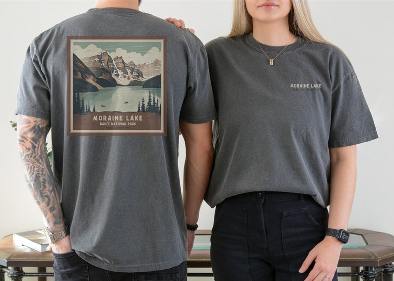 K&ouml;nnte beinhalten: Dunkelgraue T-Shirts mit einem Moraine Lake-Motiv. Auf der R&uuml;ckseite des Shirts befindet sich ein quadratisches Bild eines Sees, der von Bergen und B&auml;umen umgeben ist. Der Text "Moraine Lake Banff National Park" steht darunter. Auf der Vorderseite des Shirts steht "Moraine Lake".