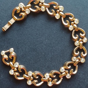 May include: A gold bracelet with a delicate, ornate design featuring a series of linked hearts. Each heart is accented with small, clear rhinestones.