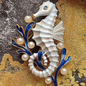 Puede incluir: Un broche de caballito de mar de esmalte blanco con detalles azules y perlados. El caballito de mar está enroscado alrededor de una rama azul y dorada con detalles perlados.