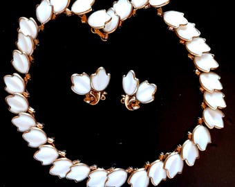 May include: A vintage jewelry set featuring a necklace and matching earrings. The set has a gold-tone metal setting with white, petal-shaped accents. The necklace and earrings are arranged on a black background.