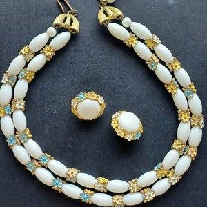 May include: A white and gold necklace with white oval beads and small blue and yellow flowers. Matching earrings with white oval stones and blue and yellow flowers.