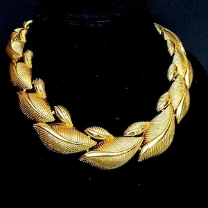 May include: A gold-toned necklace featuring a repeating leaf design. The leaves are intricately detailed with textured veins, creating a realistic appearance. The necklace is displayed against a black background, highlighting its metallic sheen.