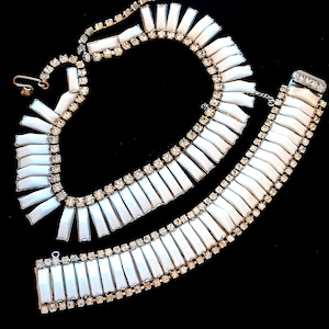 Könnte beinhalten: Ein Vintage-Set aus weißer und silberner Halskette und Armband. Der Schmuck besteht aus rechteckigen weißen Segmenten, die von kleinen, funkelnden Strasssteinen umrandet sind. Halskette und Armband werden auf schwarzem Hintergrund präsentiert, um ihr elegantes Design hervorzuheben.