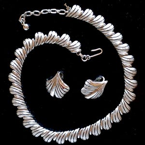 May include: A silver-toned jewelry set featuring a necklace and matching earrings. The necklace and earrings have a textured, ridged design. The necklace is a choker style with an adjustable chain. The earrings are small, leaf-shaped studs. The set is displayed against a black background.