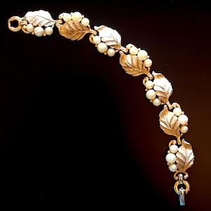 May include: Gold-toned bracelet featuring a leaf and pearl design. The bracelet has detailed leaves and clusters of small, round pearls. The clasp is a simple hook and eye closure. The jewelry is set against a black background.