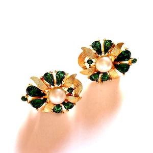 Puede incluir: Un par de pendientes dorados, cada uno con una perla central rodeada de gemas verdes esmeralda en forma de lágrima. Los pendientes tienen un diseño floral con detalles de hojas doradas texturizadas, sobre un fondo blanco.
