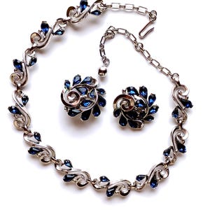 Puede incluir: Un conjunto de joyas vintage con un collar y pendientes a juego. Las piezas de metal plateado están adornadas con pedrería azul oscuro en forma de lágrima. El collar tiene un diseño decorativo y fluido.