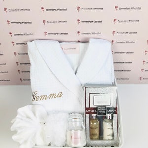 May include: A white bathrobe with the name "Gemma" embroidered on the front, a white washcloth, a white hand towel, a clear glass jar with a pink candle, a silver gift box with two bottles of bath products, and a white wicker basket.