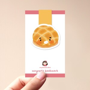 Op de afbeelding: Magnetische boekenlegger met een cartoon illustratie van een lachende, goudbruine melon pan. De boekenlegger heeft een witte achtergrond met roze en gele accenten en de tekst "magnetic bookmark".