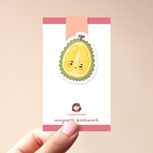 Op de afbeelding: Een roze en witte magnetische bladwijzer met een schattig cartoon durian-fruit met een glimlach. De bladwijzer heeft de tekst "raychoostudios magnetic bookmark" erop gedrukt.