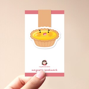 Op de afbeelding: Een roze en witte magnetische bladwijzer met een cartoon-eierkoek met een schattig gezicht. De bladwijzer heeft de tekst "raychoostudios magnetic bookmark" erop gedrukt.