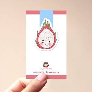 Op de afbeelding: Een roze en witte magnetische bladwijzer met een cartoon drakenvrucht met een glimlach. De bladwijzer heeft de tekst "raychoostudios magnetic bookmark" erop gedrukt.