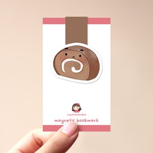 Op de afbeelding: Een bruine en witte swirl cake roll met een schattig gezicht, op een roze en witte kaart met de tekst "raychoostudios magnetic bookmark".