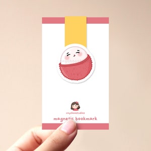 Op de afbeelding: Een witte magnetische bladwijzer met een roze en rode lychee-vrucht met een schattig cartoon-gezicht. De bladwijzer heeft een geel lint en de tekst "raychoosstudios magnetic bookmark".