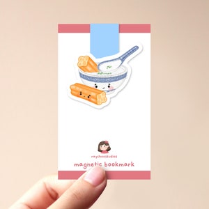 May include: A white and blue ceramic bowl with a smiling face, filled with white porridge and green onions. Two orange and yellow fried dough sticks with smiling faces are next to the bowl. The image is on a white card with the text "raychoostudios magnetic bookmark".