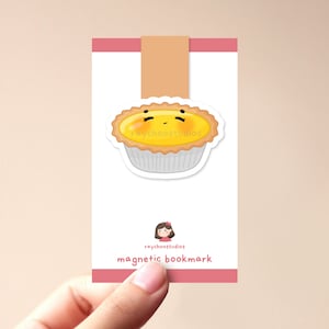 Op de afbeelding: Een roze en witte magnetische bladwijzer met een cartoon-eierkoek met een schattig gezicht. De bladwijzer heeft de tekst "raychoosstudios magnetic bookmark" erop gedrukt.
