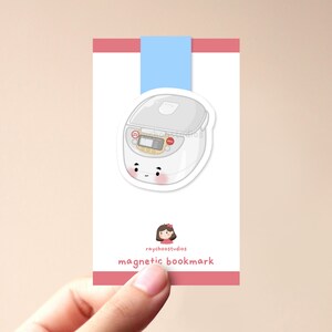 Op de afbeelding: Een witte en grijze cartoon rijstkoker met een rode knop en een glimlachend gezicht. De rijstkoker staat op een roze en witte kaart met de tekst "raychoostudios magnetic bookmark".
