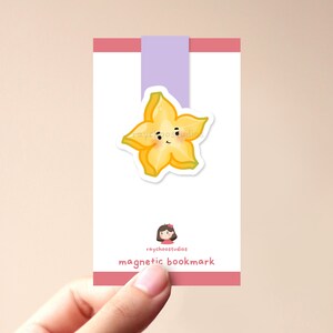 Op de afbeelding: Een roze en witte magnetische bladwijzer met een gele stervrucht met een schattig gezicht. De bladwijzer heeft de tekst "raychoosstudios" en "magnetische bladwijzer".