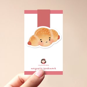 Op de afbeelding: Een roze en witte kaart met een cartoonillustratie van een croissant met een glimlachend gezicht. De tekst "raychoostudios magnetic bookmark" staat op de kaart gedrukt.