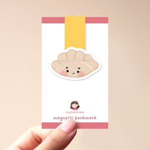 以下が含まれることがあります： 笑顔の漫画の餃子が描かれたピンクと白の磁気ブックマーク。ブックマークには「raychoostudios magnetic bookmark」というテキストが印刷されています。