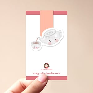 Könnte beinhalten: Ein rosa-weißes magnetisches Lesezeichen mit einer Cartoon-Teekanne, die Tee in eine Tasse gießt. Die Teekanne und die Tasse haben süße Gesichter. Das Lesezeichen trägt den Text "raychoostudios magnetic bookmark".