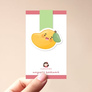 Op de afbeelding: Een roze en witte kaart met een cartoon mango met een glimlachend gezicht en een groen blad. Op de kaart staat onderaan "raychoostudios magnetic bookmark".