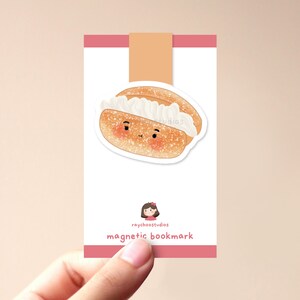 Op de afbeelding: Een roze en witte magnetische bladwijzer met een cartoonillustratie van een roomsoezen met een glimlach. De bladwijzer heeft de tekst "raychoostudios magnetic bookmark" erop gedrukt.