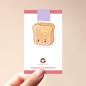 Op de afbeelding: Een schattige cartoonillustratie van een snee toast met een klontje boter erop. De toast heeft een glimlachend gezicht en de boter heeft een knipperend oog. De illustratie is gedrukt op een roze en witte magnetische bladwijzer met de tekst "raychoostudios magnetic bookmark".