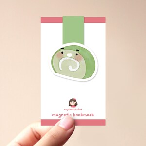 Op de afbeelding: Groen en wit cartoon rolcake-bladwijzer met een schattig gezicht. De bladwijzer is magnetisch en heeft de tekst "raychoostudios magnetic bookmark" onderaan.