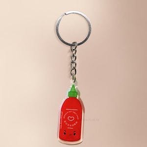May include: A red Sriracha hot sauce bottle keyring with a silver ring and chain. The keyring features a green cap, a heart, and text in English and another language. The bottle has a smiling face.