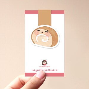 Op de afbeelding: Een schattige cartoonrolcake met een glimlachend gezicht. De rolcake is bruin met een witte vulling. De afbeelding heeft de tekst "raychoostudios magnetic bookmark" onderaan.