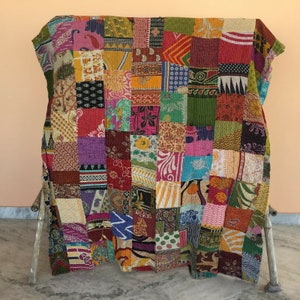 May include: A colorful patchwork quilt made from a variety of fabrics. The quilt is made up of squares of different colors and patterns, including floral, paisley, and geometric designs. The quilt is folded and displayed on a wooden stand.