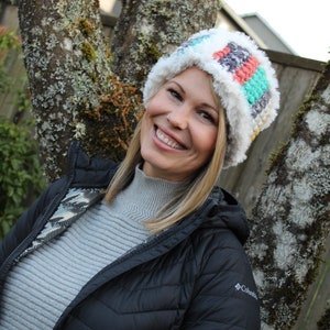May include: A woman wearing a black jacket with a white fur trim and a colorful crocheted hat. The hat has stripes of white, gray, pink, teal, and yellow.