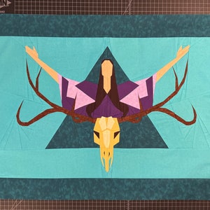 Puede incluir: Un tapiz de pared de tela azul turquesa con un diseño geométrico que presenta a una mujer con los brazos extendidos, una calavera de ciervo y astas. El diseño está centrado en una forma triangular.