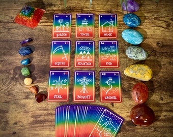 May include: A set of 10 tarot cards with rainbow backgrounds and white illustrations. The cards are laid out in a grid pattern on a wooden surface. There are also several colorful stones scattered around the cards.