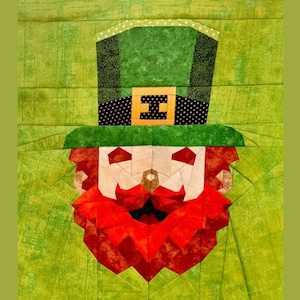 Op de afbeelding: Een groene leprechaun met een rode baard en een groene hoed met een gouden gesp en zwarte stippen. De hoed heeft een witte rand met witte stippen. De leprechaun is gemaakt van stof en heeft een patchwork-ontwerp.