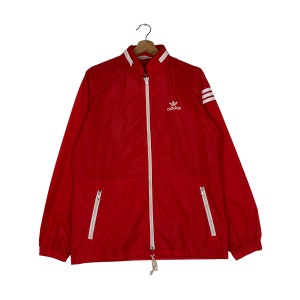 Può includere: Una giacca a vento rossa con strisce bianche sulle maniche e un logo Adidas bianco sul petto.
