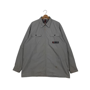 May include: A light gray jacket with a collared neckline and a full-length zipper. The jacket features two chest pockets with button closures and a small logo patch on the right side. The long sleeves have buttoned cuffs.