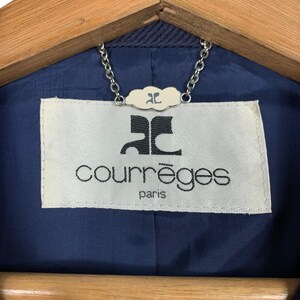 May include: Close-up of a navy blue jacket lining with a white label. The label features the brand name "courr&egrave;ges" in a sans-serif font, with "paris" below. A silver chain and a small cloud-shaped tag are attached.