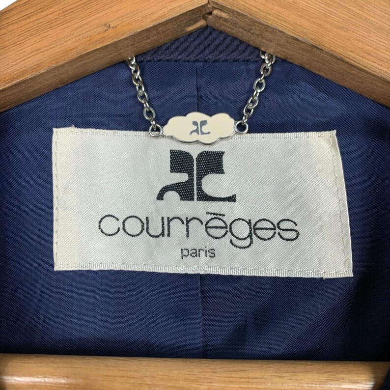 May include: Close-up of a navy blue jacket lining with a white label. The label features the brand name "courr&egrave;ges" in a sans-serif font, with "paris" below. A silver chain and a small cloud-shaped tag are attached.