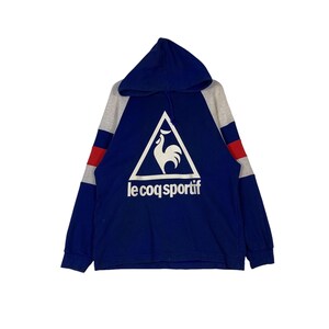 Puede incluir: Una sudadera con capucha azul, gris y roja con un logotipo de gallo blanco en un tri&aacute;ngulo y el texto "le coq sportif" debajo.
