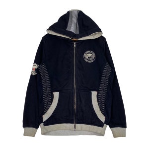 May include: Navy blue zip-up hoodie with a gray hood lining and trim. Features a circular patch with text and a stitched design on the sleeves. The hoodie has a full-length zipper and two front pockets.