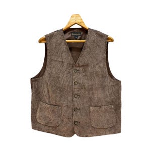 Puede incluir: Un chaleco de tweed marrón con cierre de botones y dos bolsillos. El chaleco tiene un patrón de cuadros marrones y blancos.