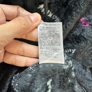 Może przedstawiać: Zbliżenie etykiety z japońskim tekstem i napisem "MADE IN CHINA". Etykieta jest przymocowana do ciemnoszarej, puszystej tkaniny z białymi i r&oacute;żowymi akcentami. Tkanina wydaje się być pluszowym materiałem.