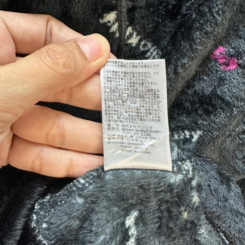 Może przedstawiać: Zbliżenie etykiety z japońskim tekstem i napisem "MADE IN CHINA". Etykieta jest przymocowana do ciemnoszarej, puszystej tkaniny z białymi i r&oacute;żowymi akcentami. Tkanina wydaje się być pluszowym materiałem.