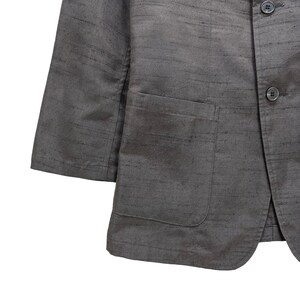 Puede incluir: Una chaqueta gris con un solo bolsillo en el pecho y dos botones. La chaqueta est&aacute; hecha de una tela texturizada.